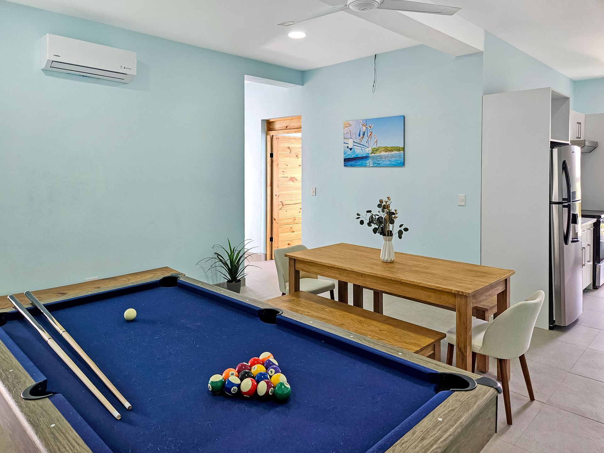 Pool table in a room with a dining table, chairs, and a kitchen area. Blue walls, light wood furniture.