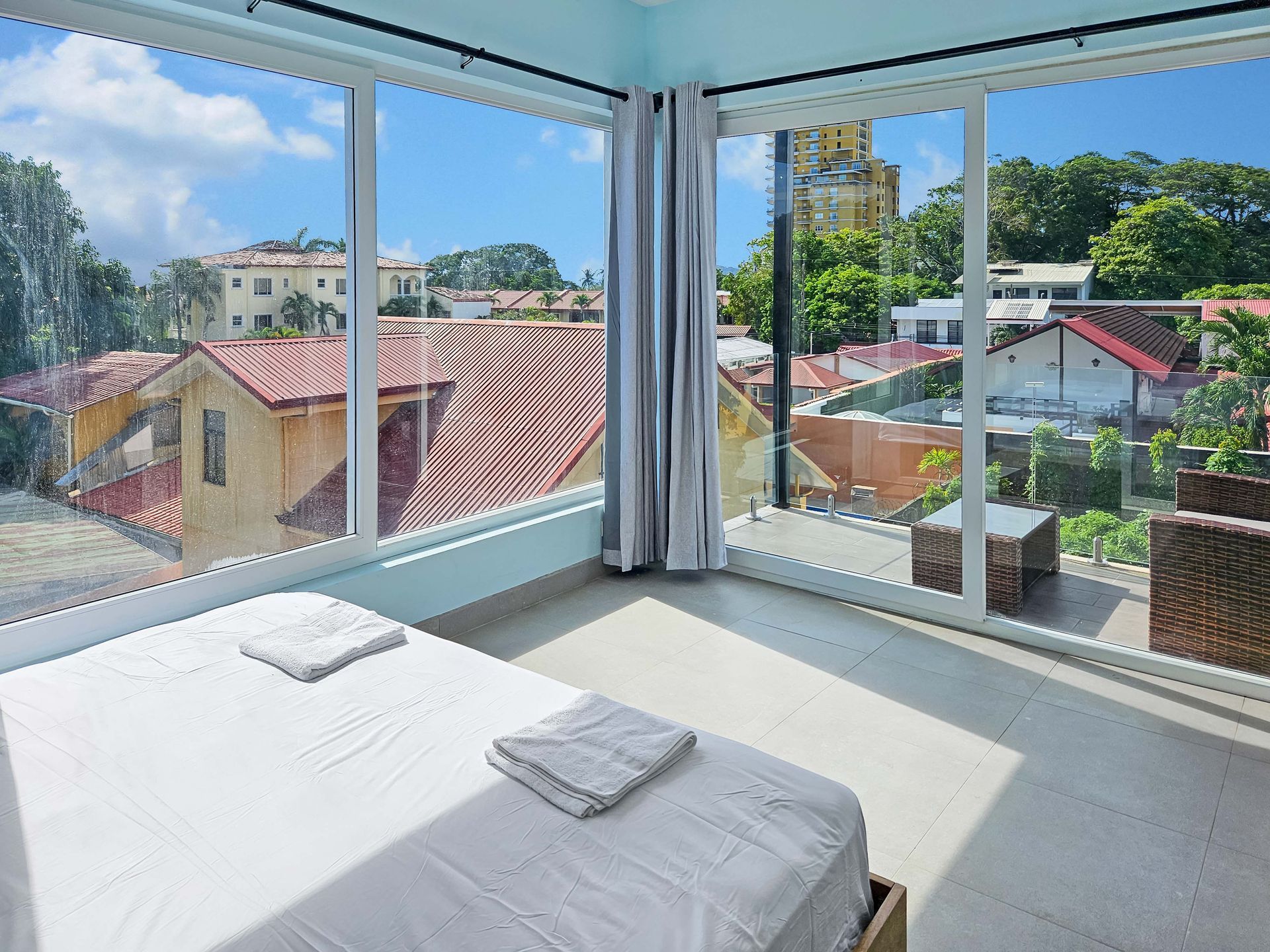 Bedroom with large windows overlooking a city, featuring a bed, and outdoor seating.