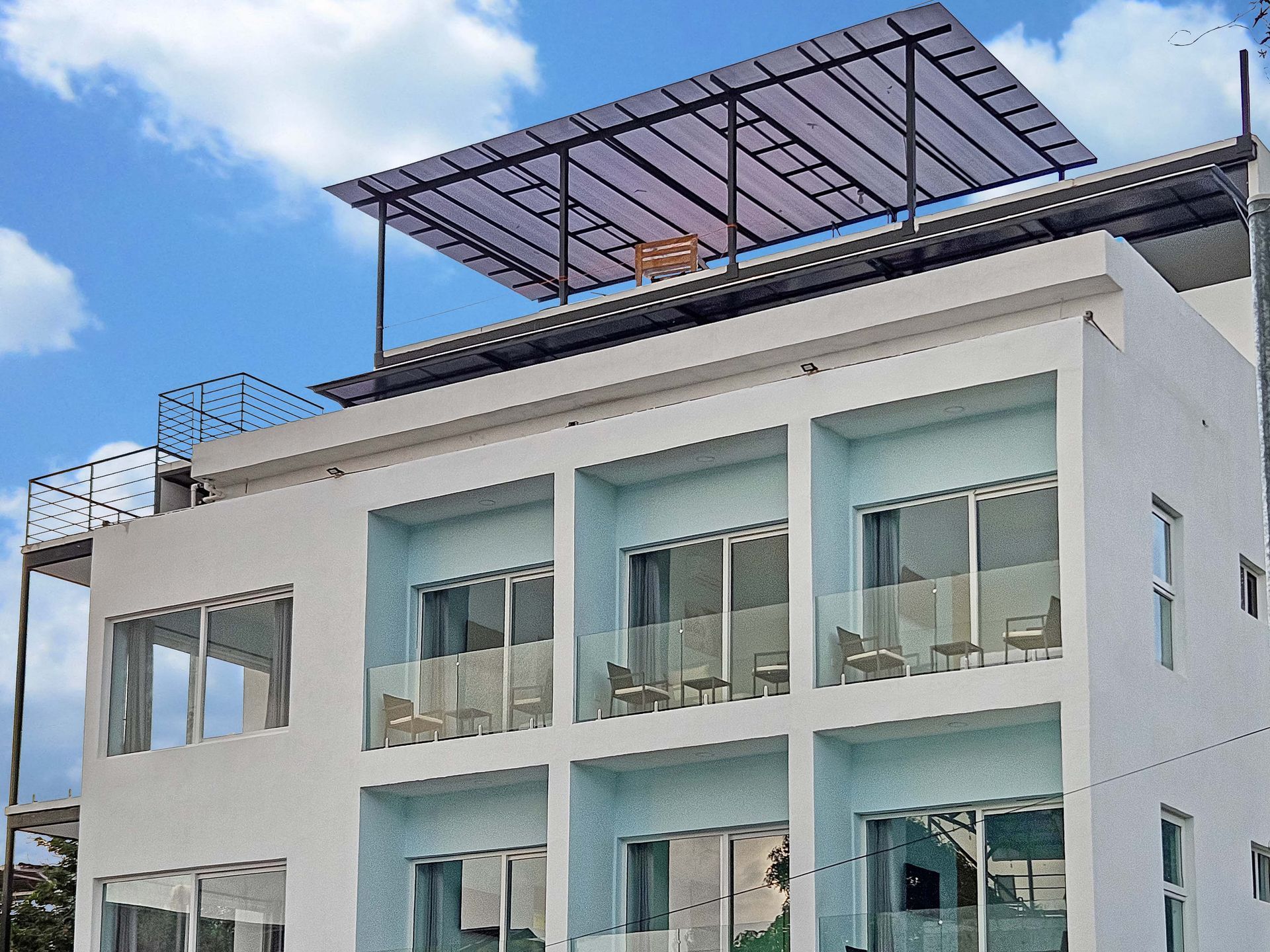 White building with balconies, glass railings, and a solar panel covered roof against a blue sky.