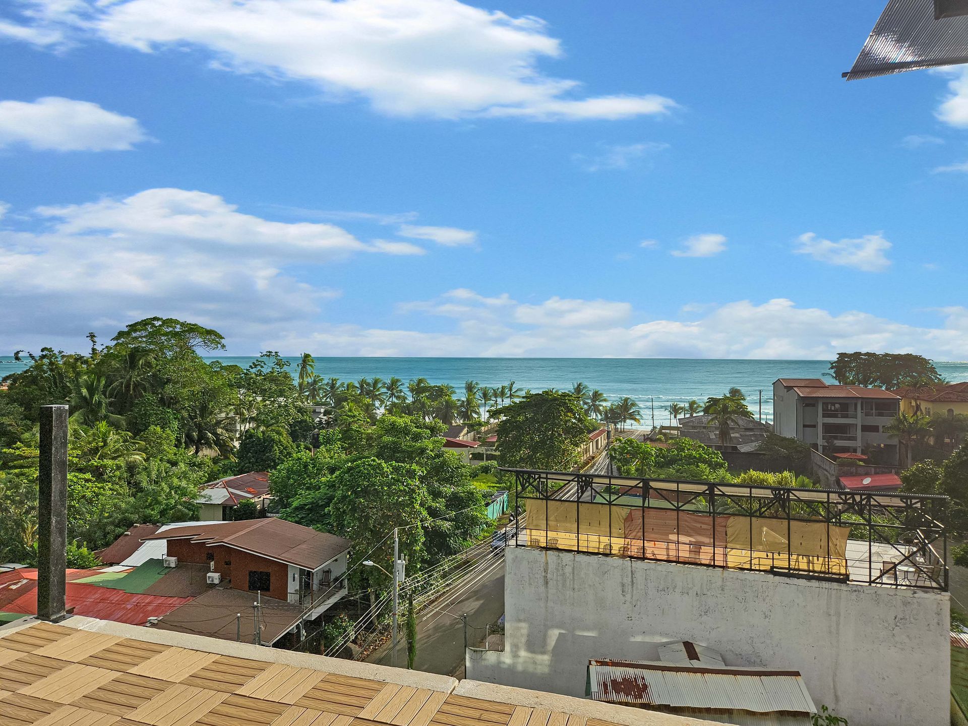 View of the ocean from a rooftop, with buildings and trees in the foreground, blue sky with clouds.