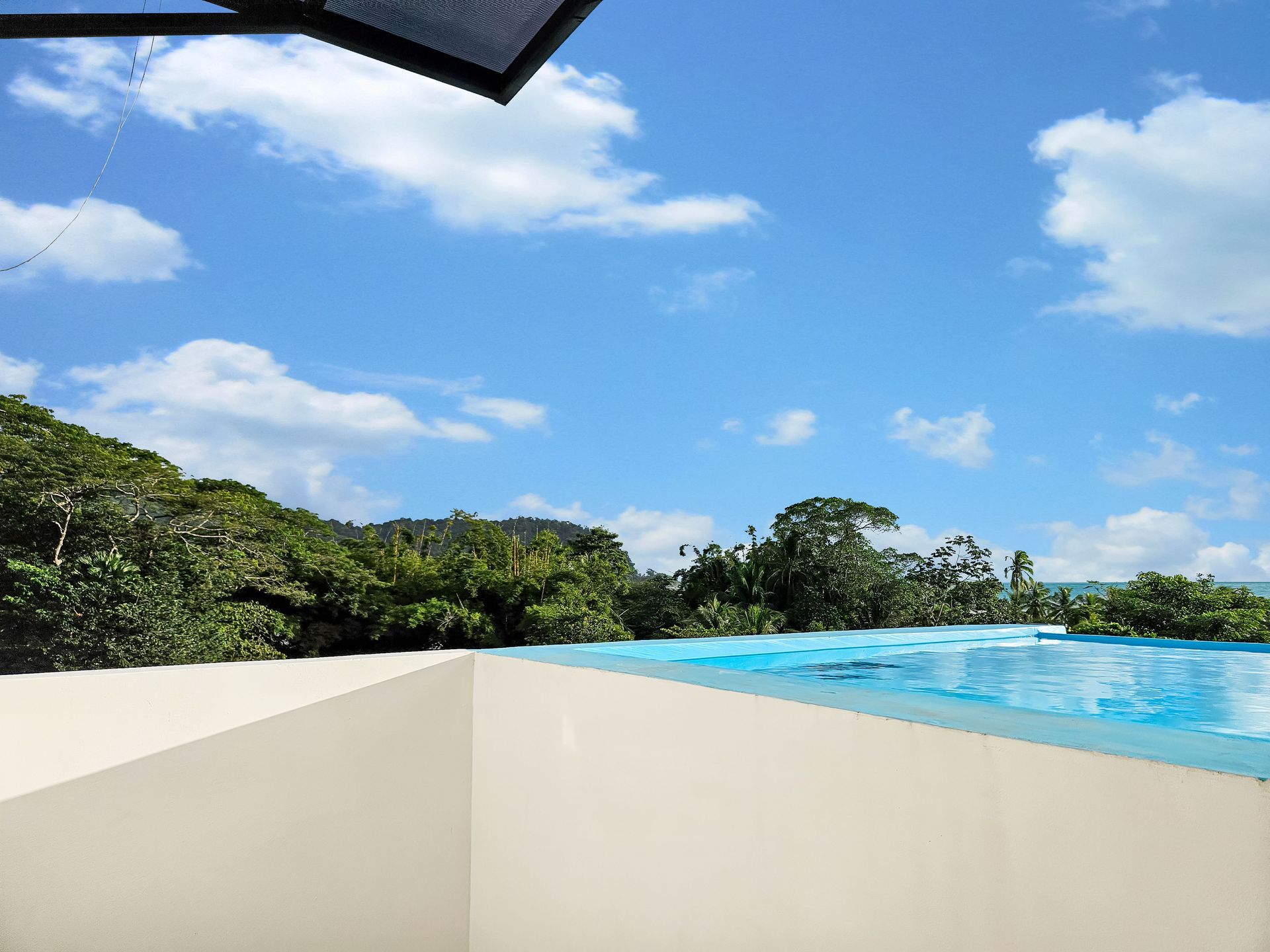 Infinity pool overlooking ocean and lush green trees under blue sky with clouds.