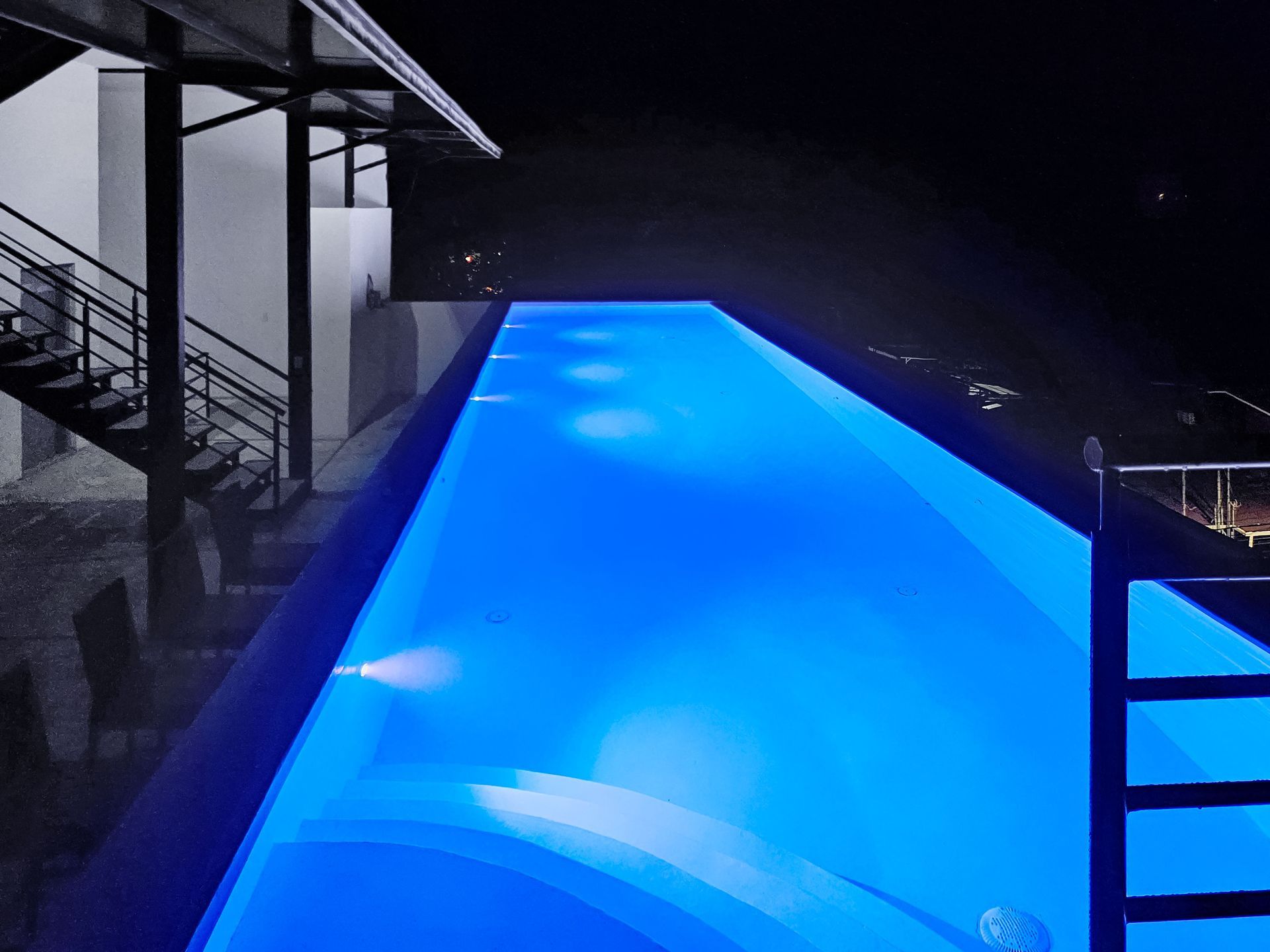 Night view of an illuminated blue infinity pool. Stairs and building in the background.