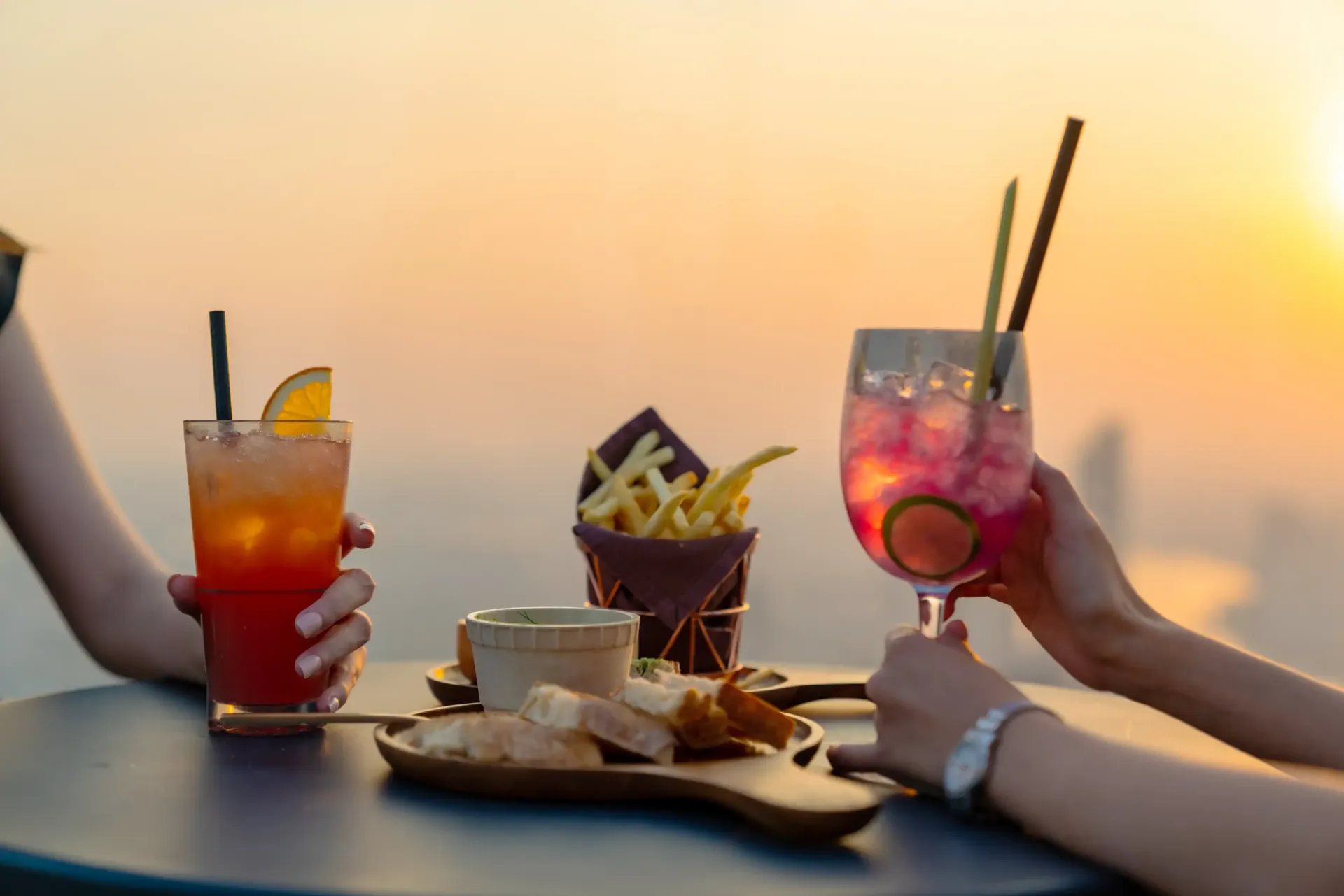 Two drinks, fries, and bread on a table at sunset. Hands holding the cocktails.