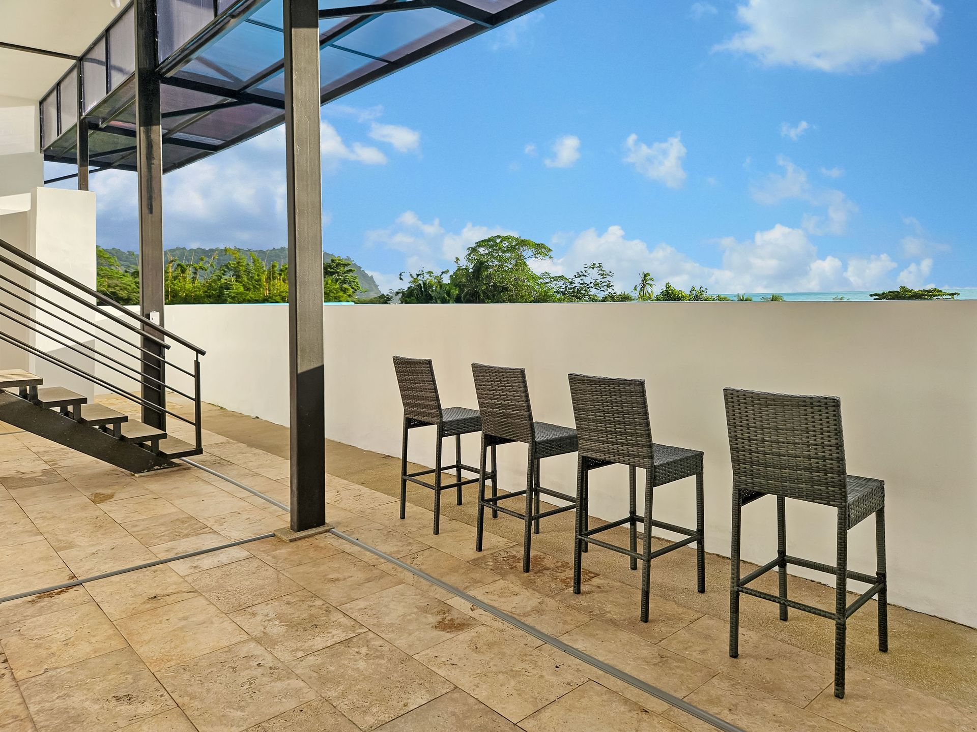 Outdoor patio with bar stools, stone floor, metal staircase, white wall, blue sky.