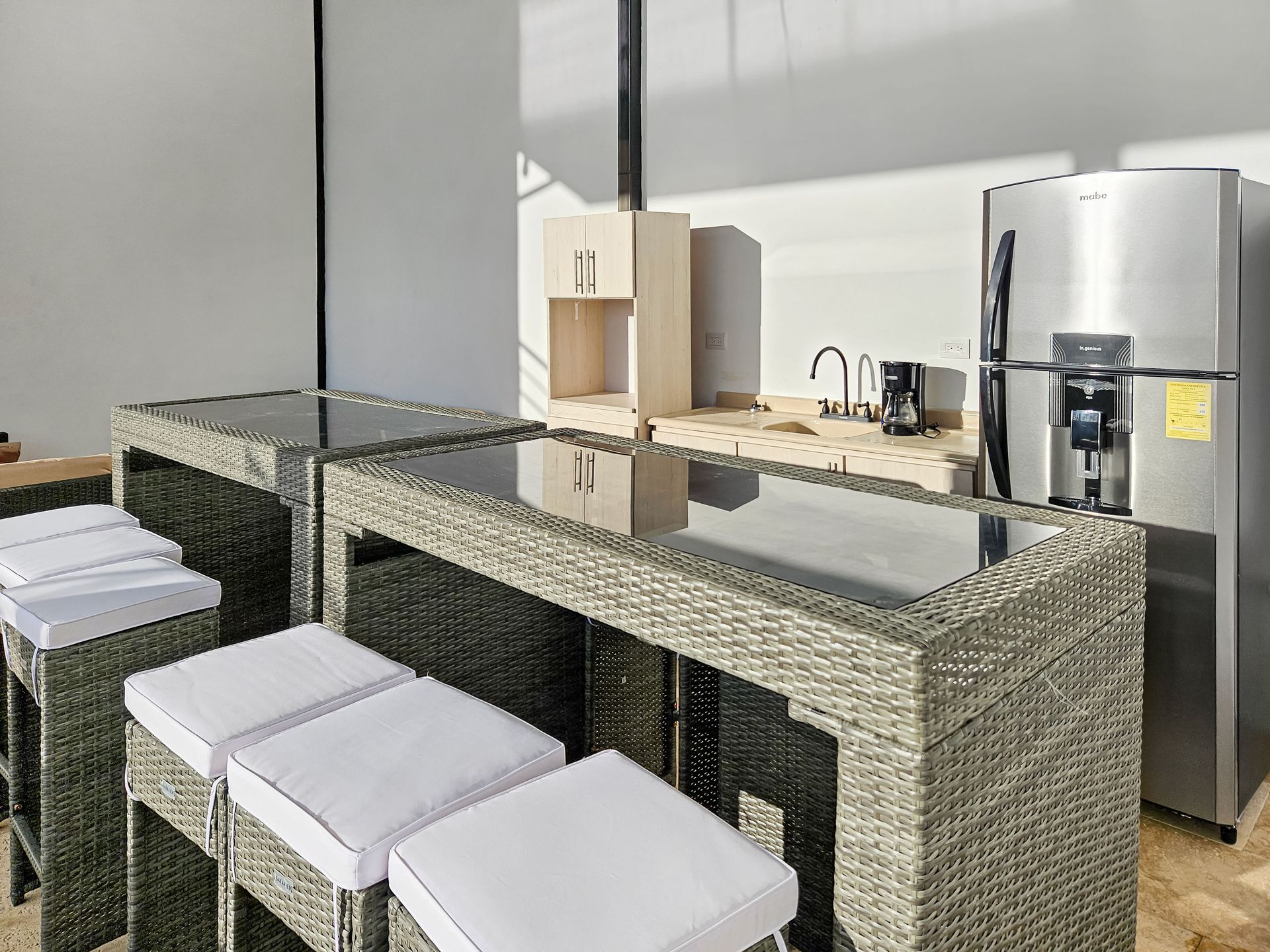 Outdoor kitchen area with woven gray bar stools and tables, stainless steel refrigerator, and light wood cabinetry.