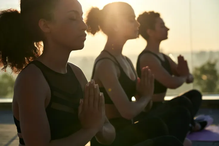 Three people meditating outdoors with hands together, backlit by the sun.