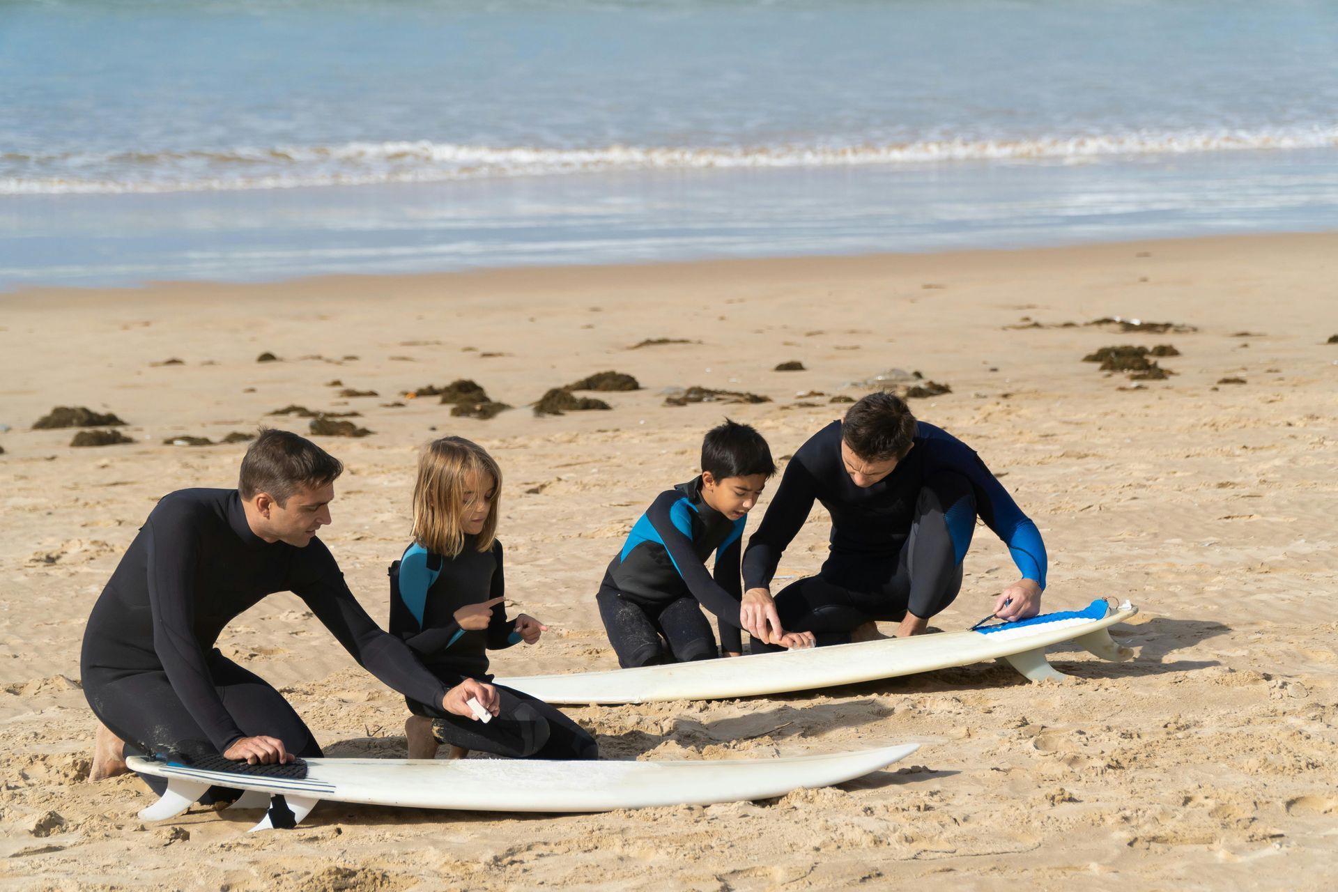 Four people kneeling on a beach, preparing surfboards. Two adults and two children in wetsuits.