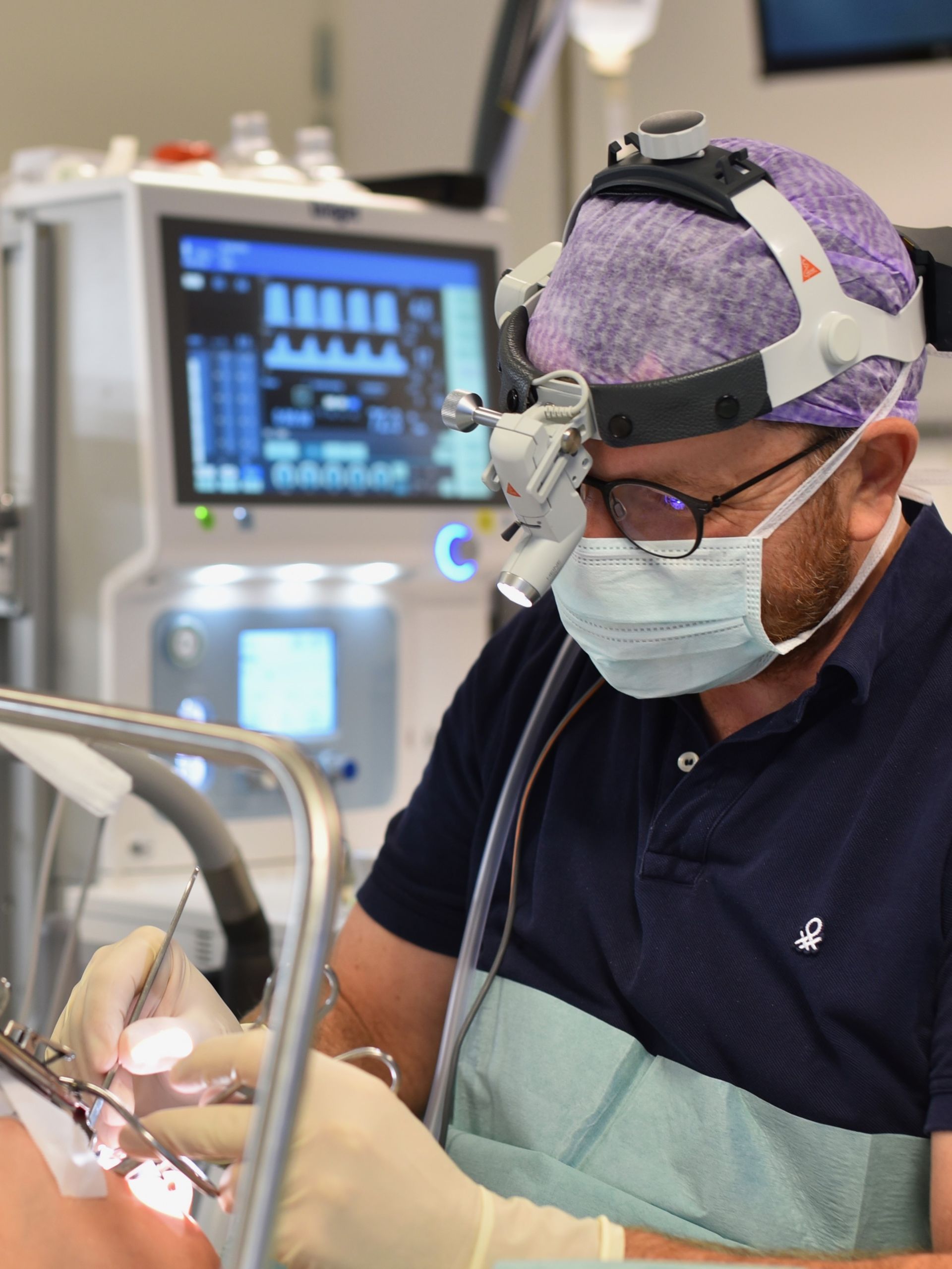 Kirurg i scrubs och pannlampa arbetar med instrument. Medicinsk utrustning i bakgrunden.
