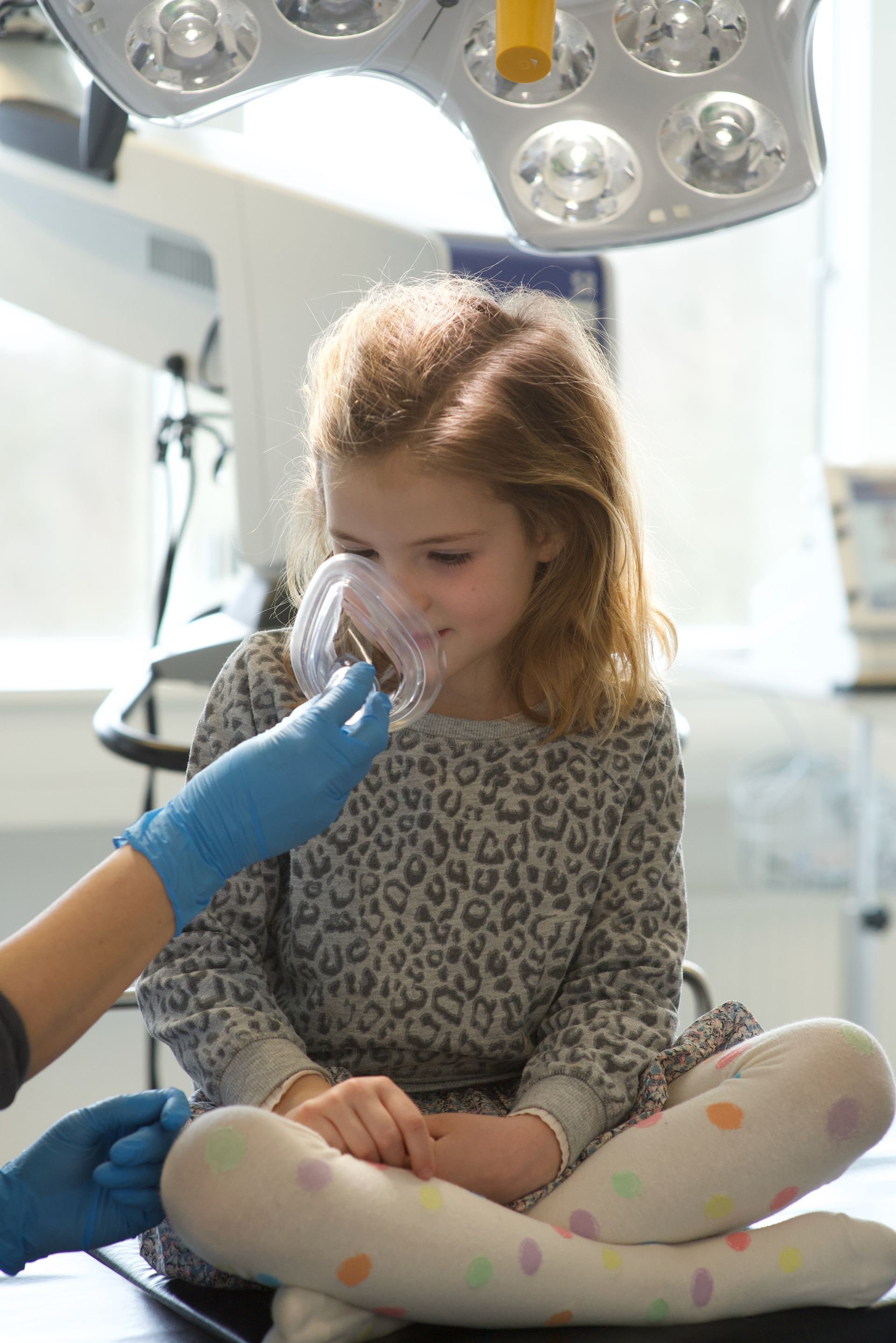 Flicka med syrgasmask; medicinsk miljö; handskbeklädd hand; leende.