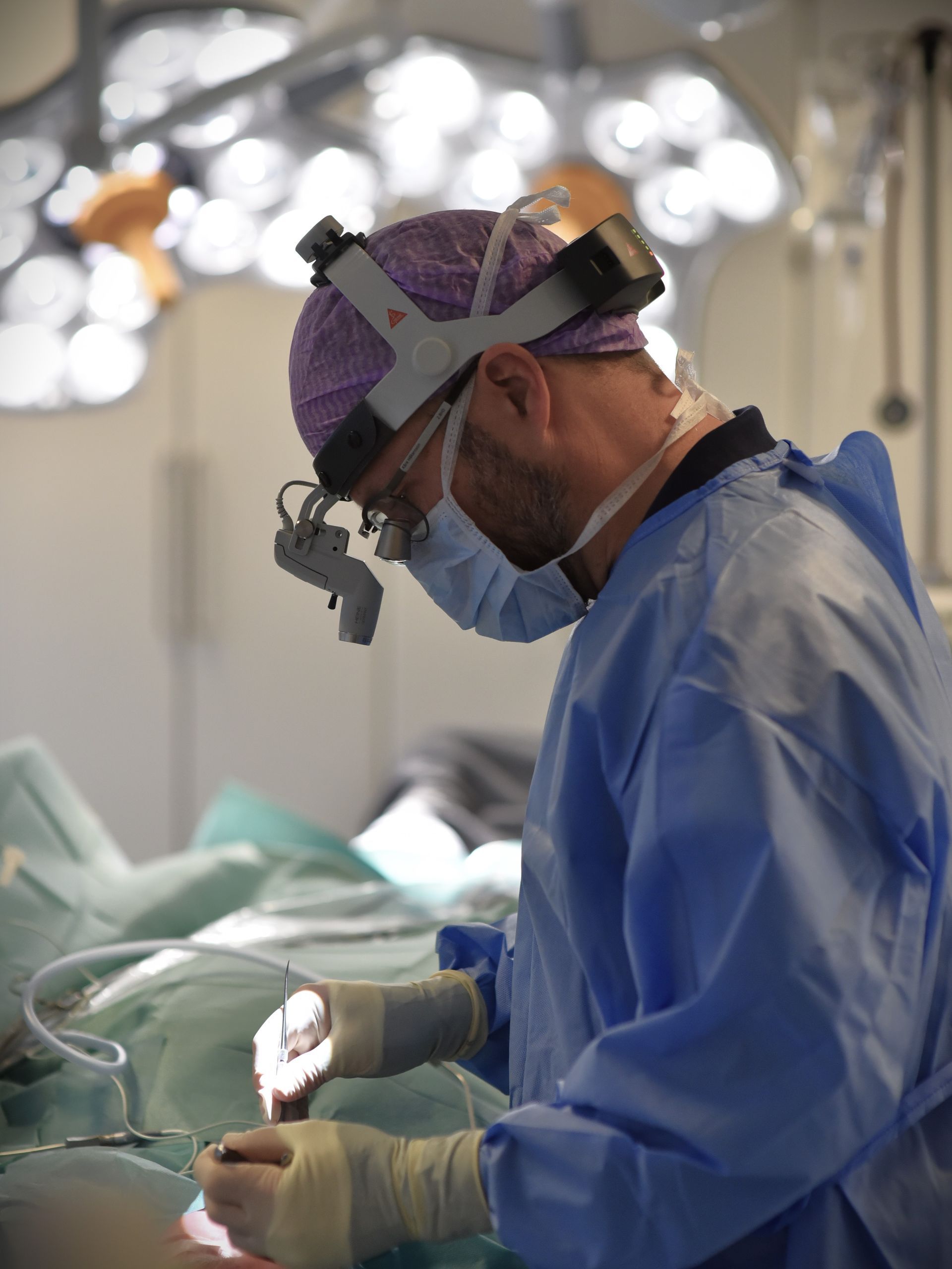 Kirurg i blå scrubs, med pannlampa och mask, opererar i en väl upplyst operationssal.