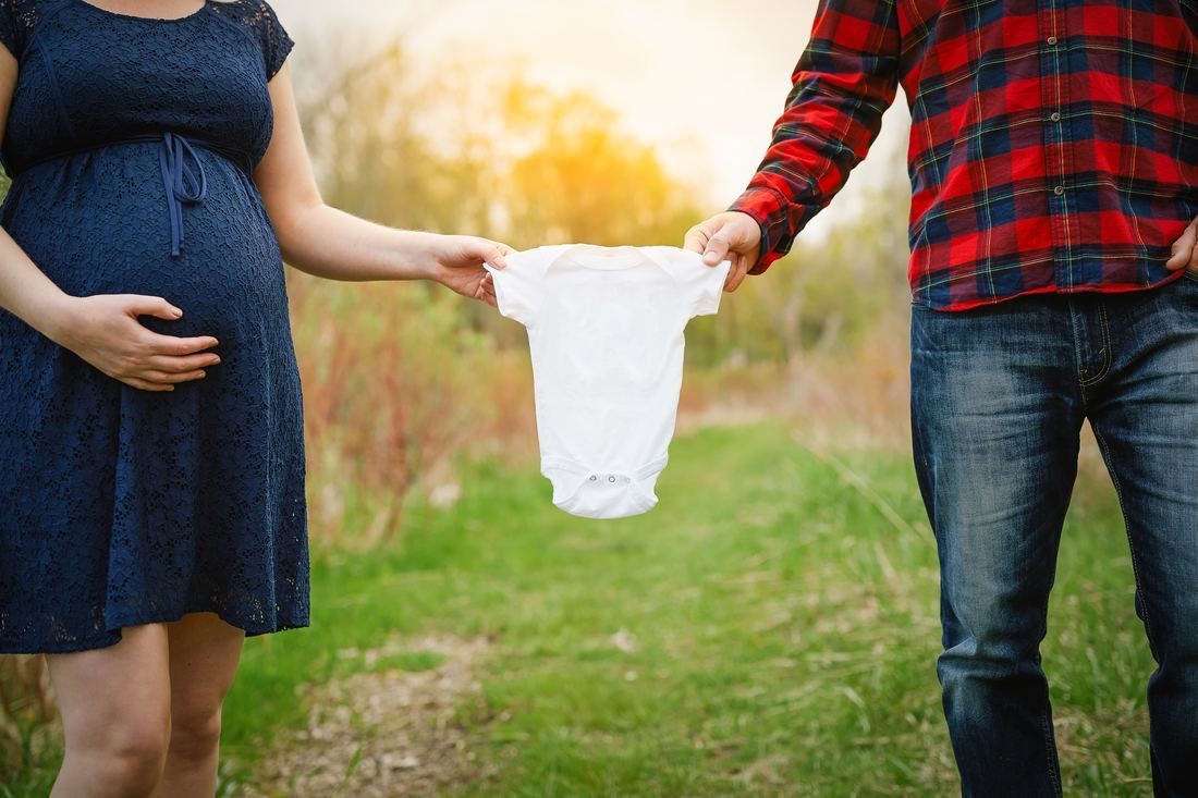 A pregnant woman and a man are holding a baby clothes in a field.