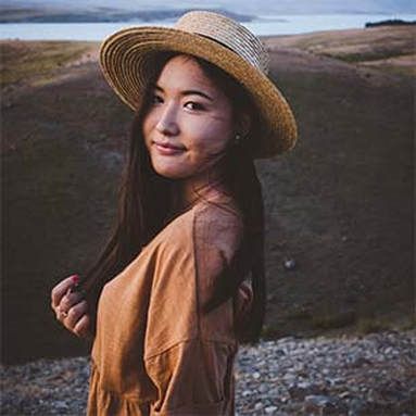 A woman wearing a straw hat and a brown dress is standing in front of a lake.