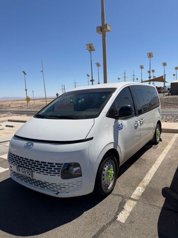 Un profesional sonriente con uniforme azul señala un autobús blanco estacionado con rayas azules.