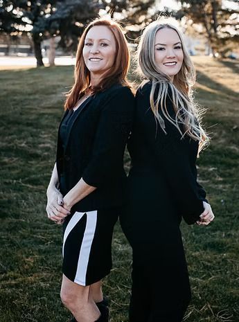 Two women in black outfits stand back-to-back on a grassy lawn, smiling.