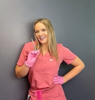 Woman in pink scrubs holds a syringe, smiling. Wearing pink gloves, against a gray wall.