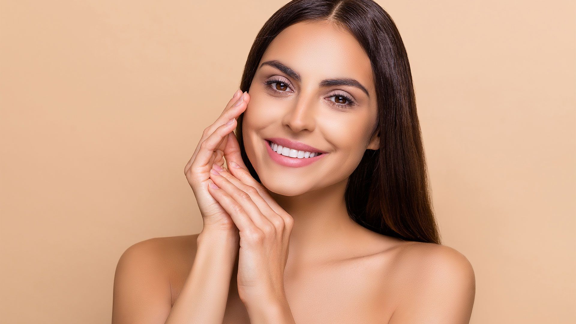 Woman with long brown hair smiles, touching face with hands. Tan background.