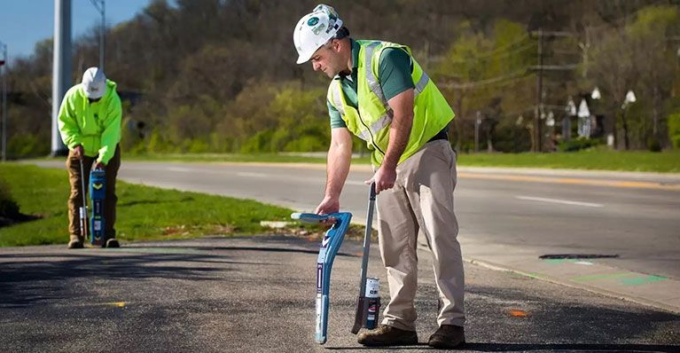 Two workers using locating equipment on a road. One kneels, one stands, both wearing safety vests.
