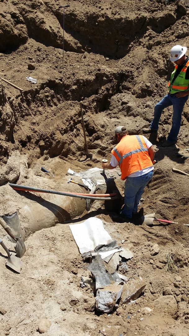 Construction workers excavating dirt, inspecting concrete structures. One wears an orange vest, one a hard hat.