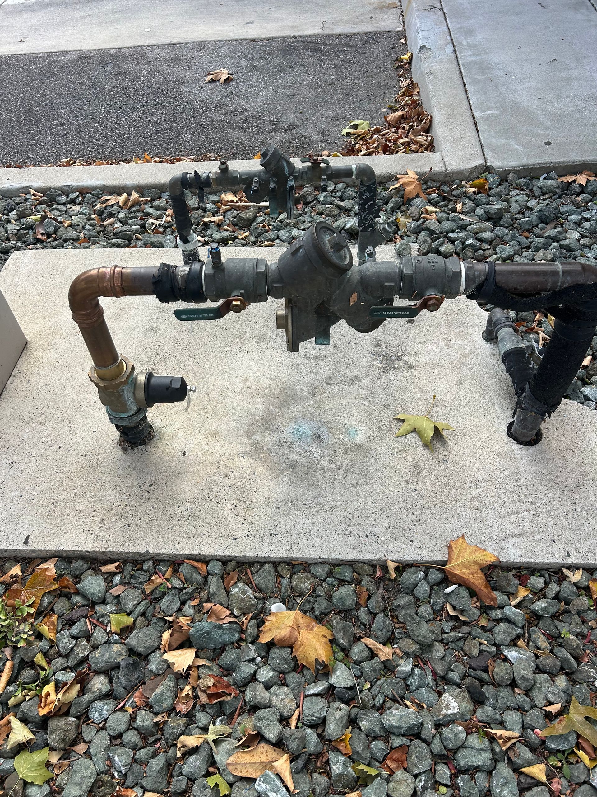 Pipes and valves on a concrete pad with gravel surroundings. Some leaves present.