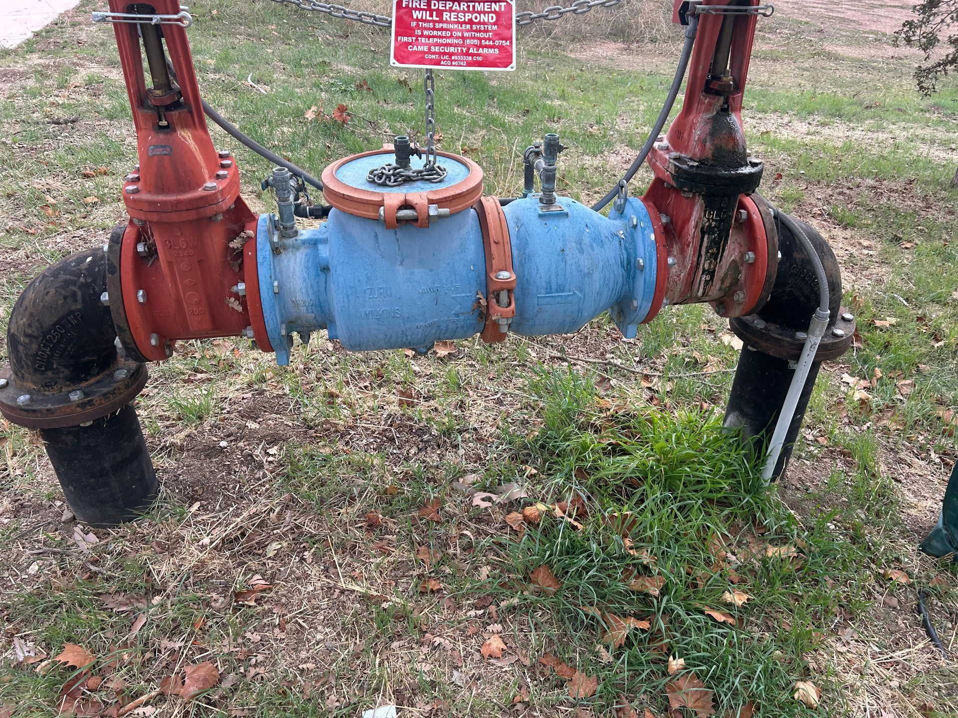 Blue and red water meter with pipes, in a grassy area, with a warning sign.