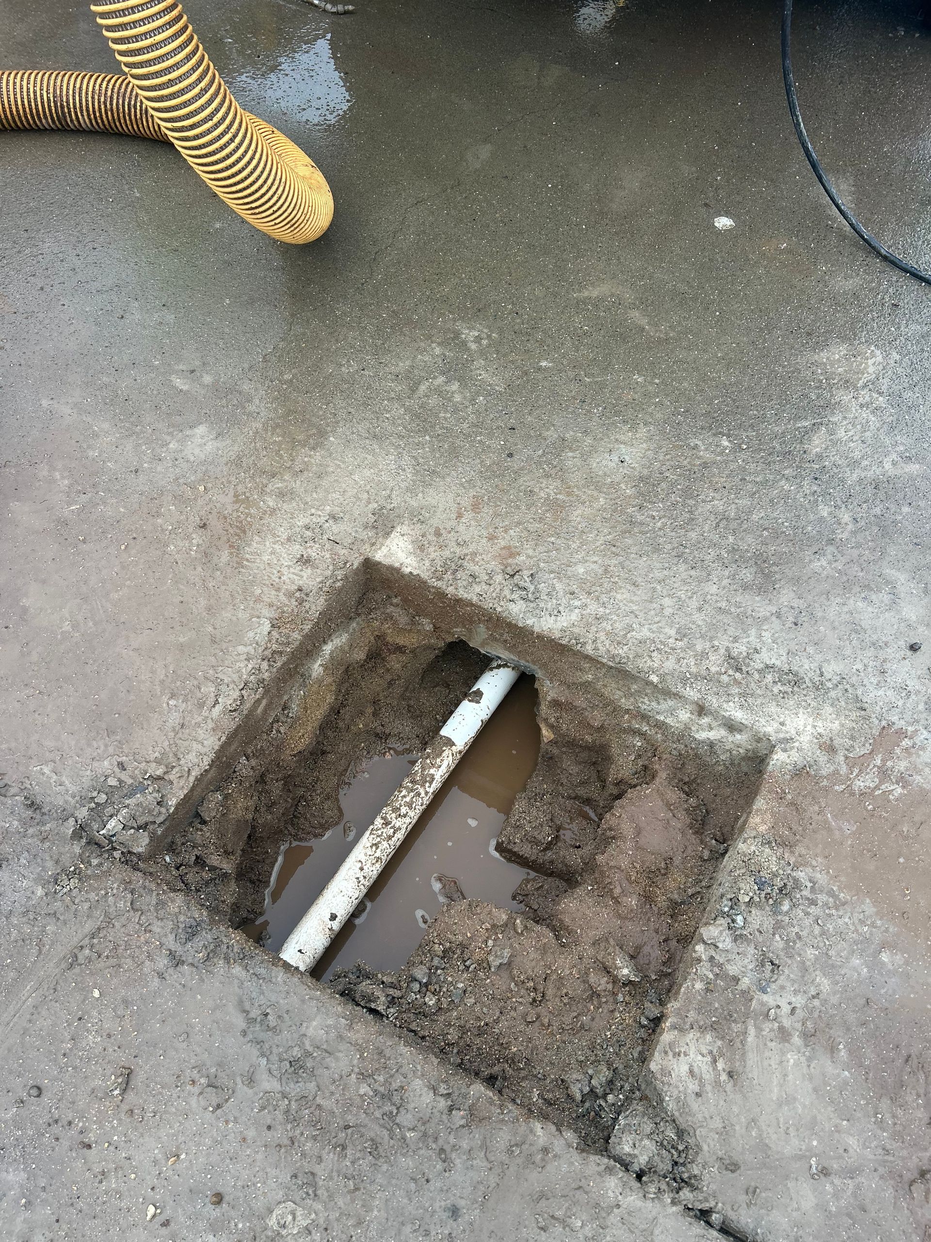A square hole in concrete reveals a white pipe, surrounded by dirt and water; a coiled hose is in the corner.