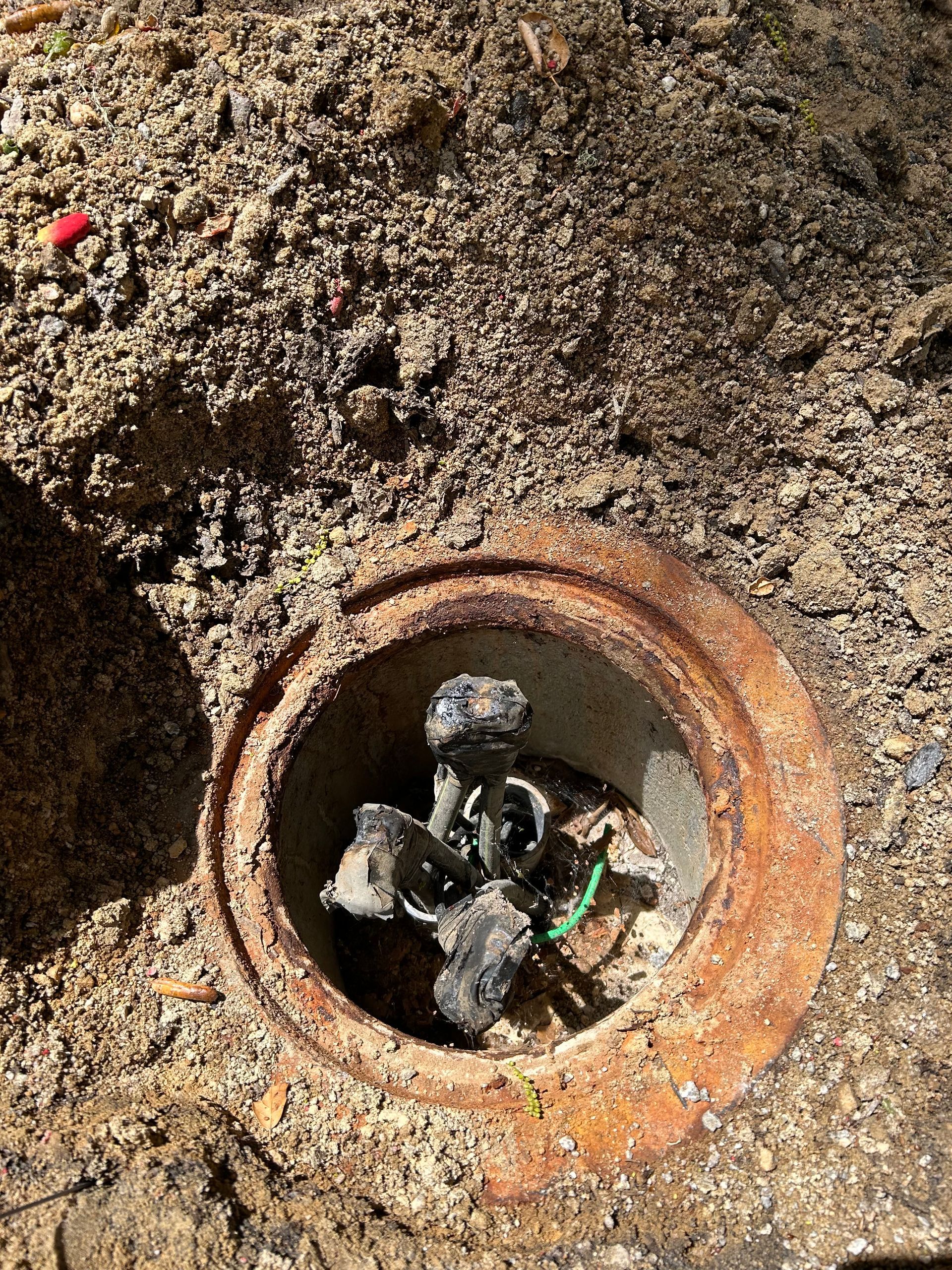 Rusty metal access cover open in dirt, revealing charred electrical wiring.