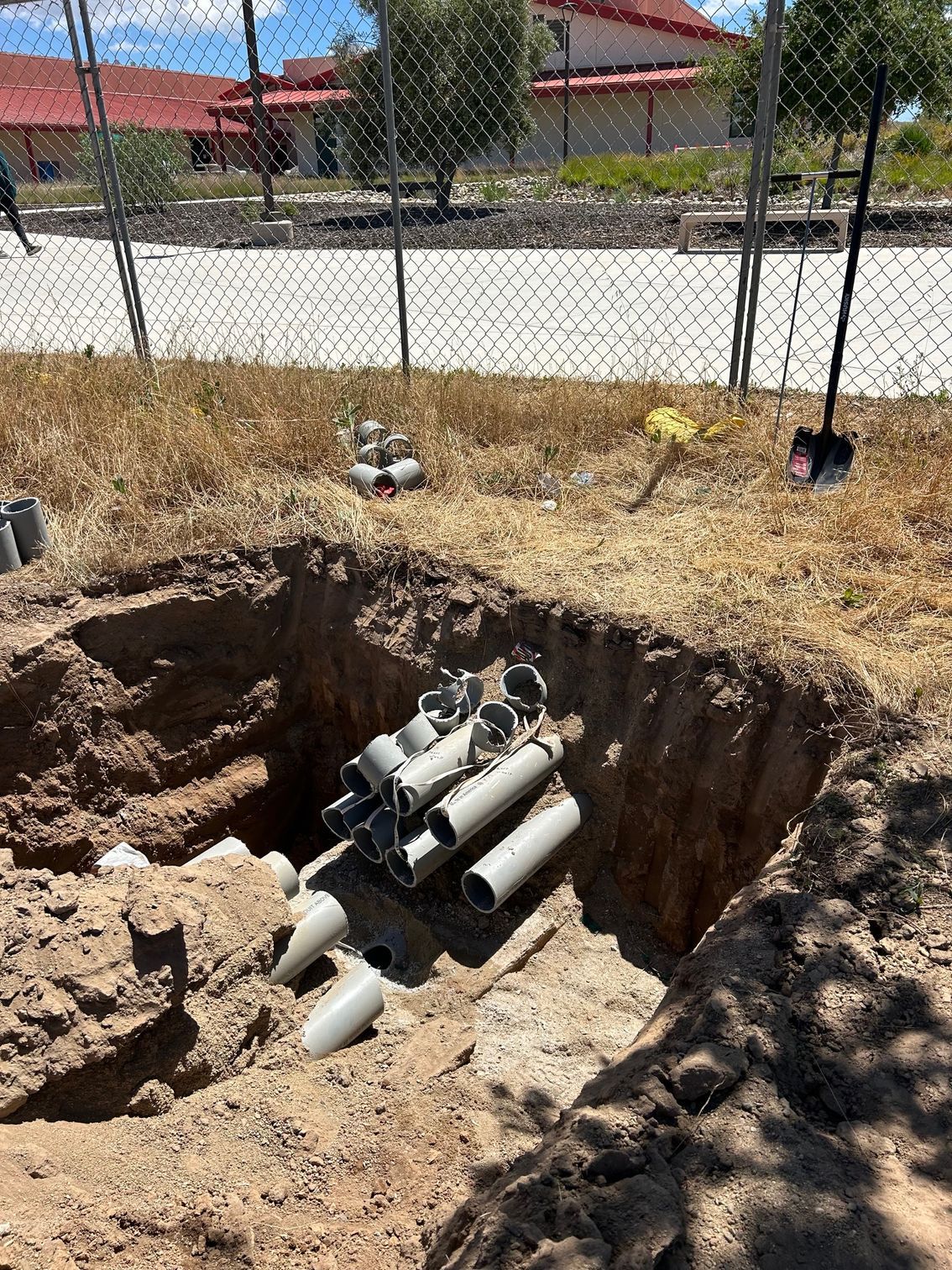 Hole dug in dirt with gray pipes inside, near a fence and building on a sunny day.