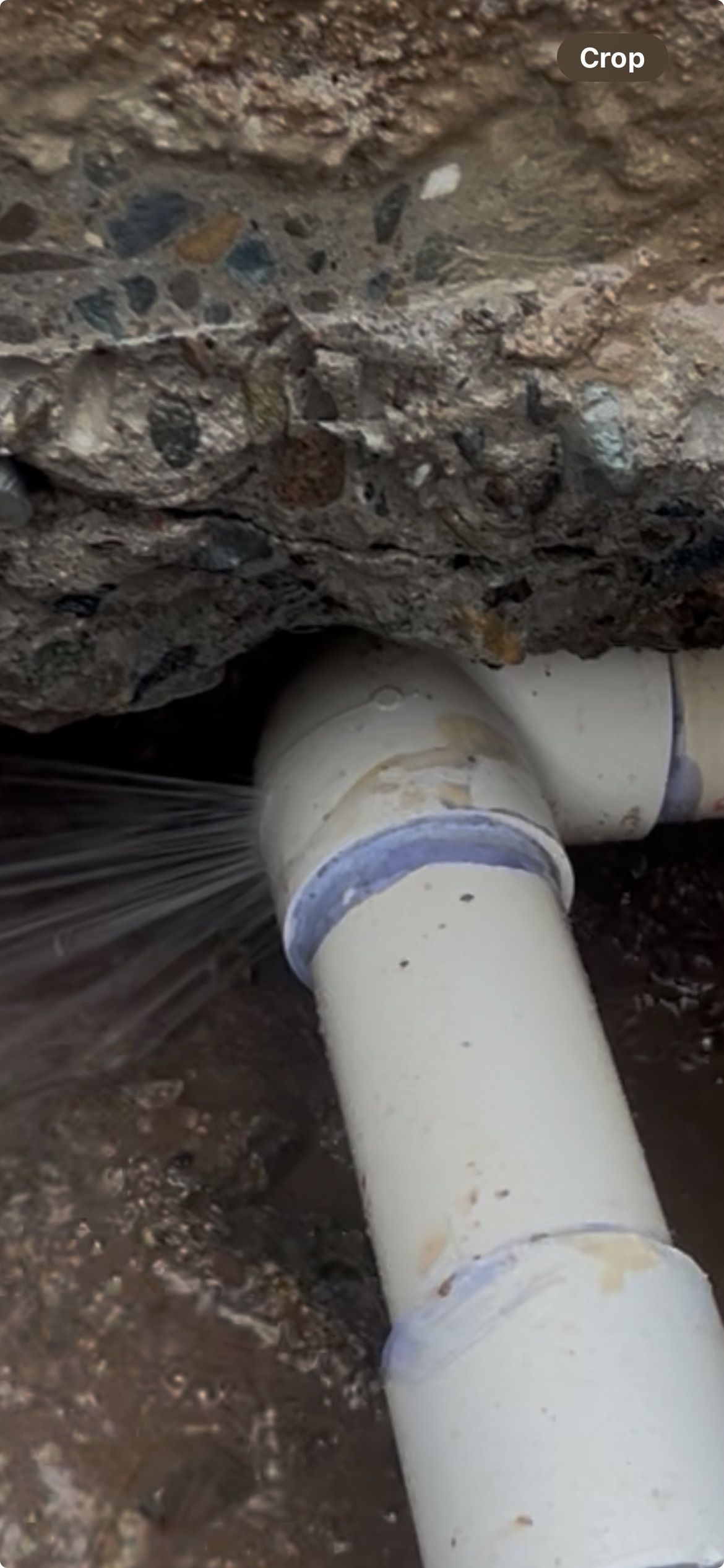 Water spurts from a broken PVC pipe joint beneath a concrete structure.