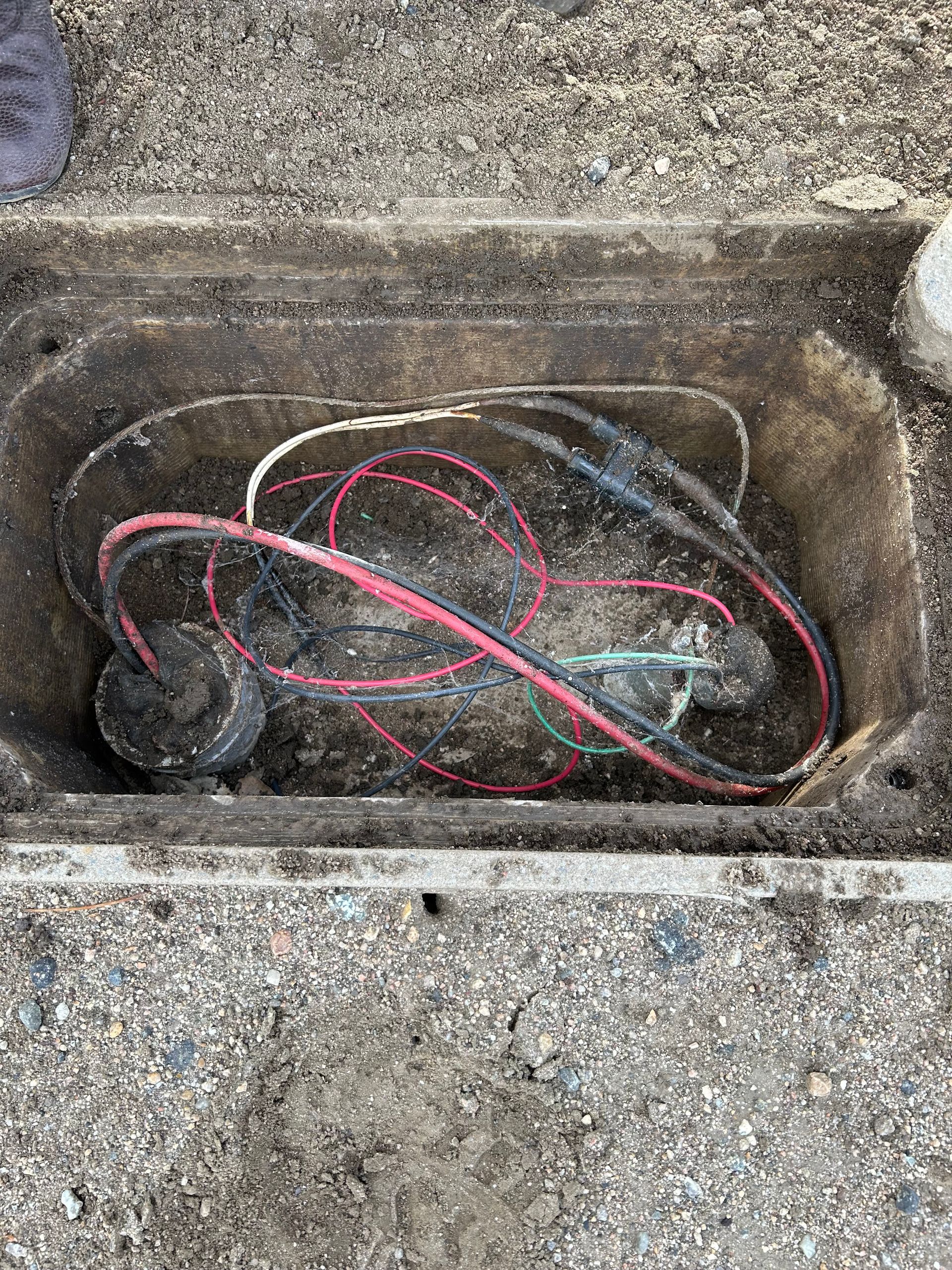 Open utility box with tangled red, black, and green wires inside. Gray dirt surrounds the rectangular opening.
