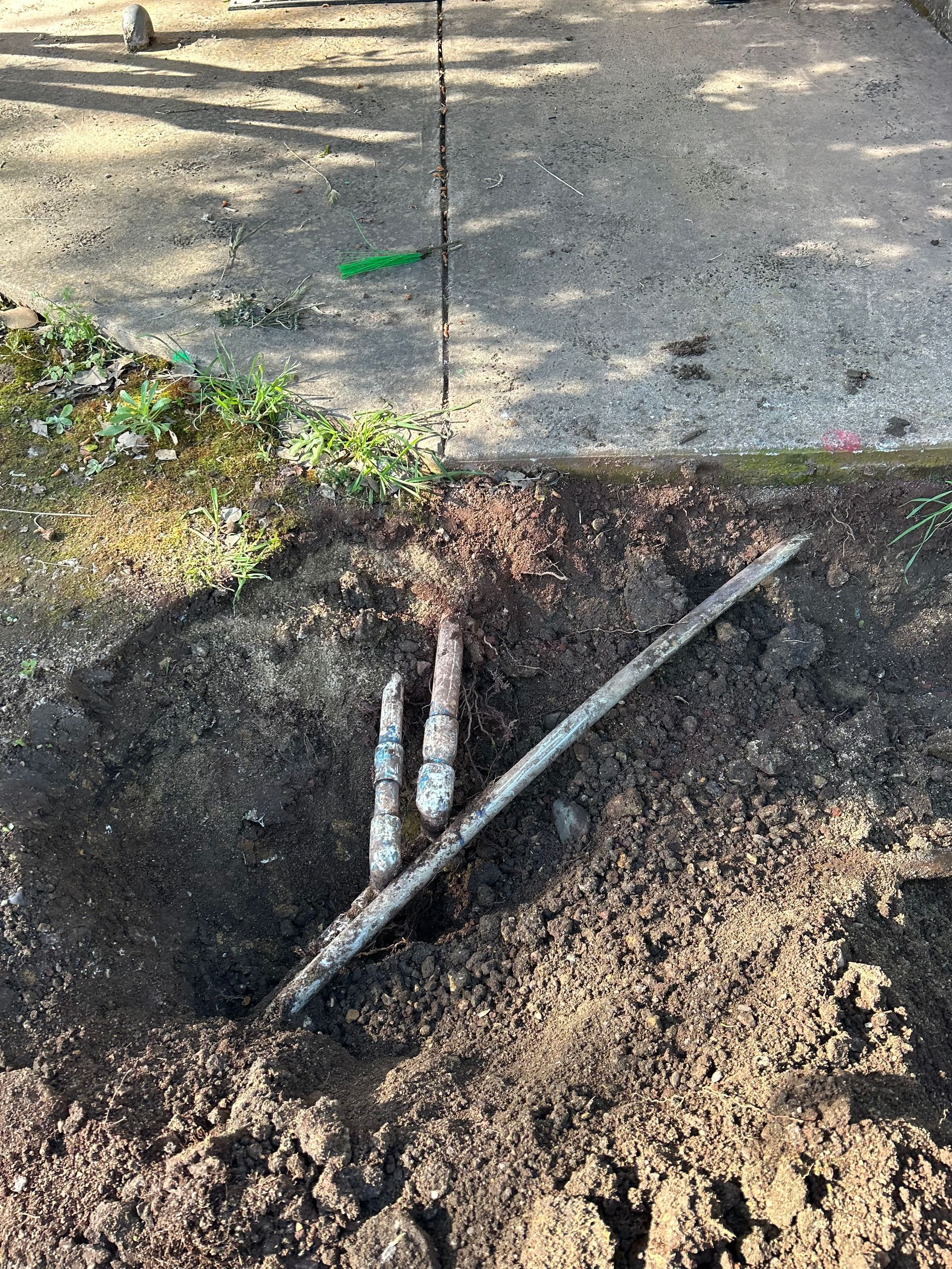 Hole dug in dirt exposing buried pipes. A concrete surface and greenery are in the background.