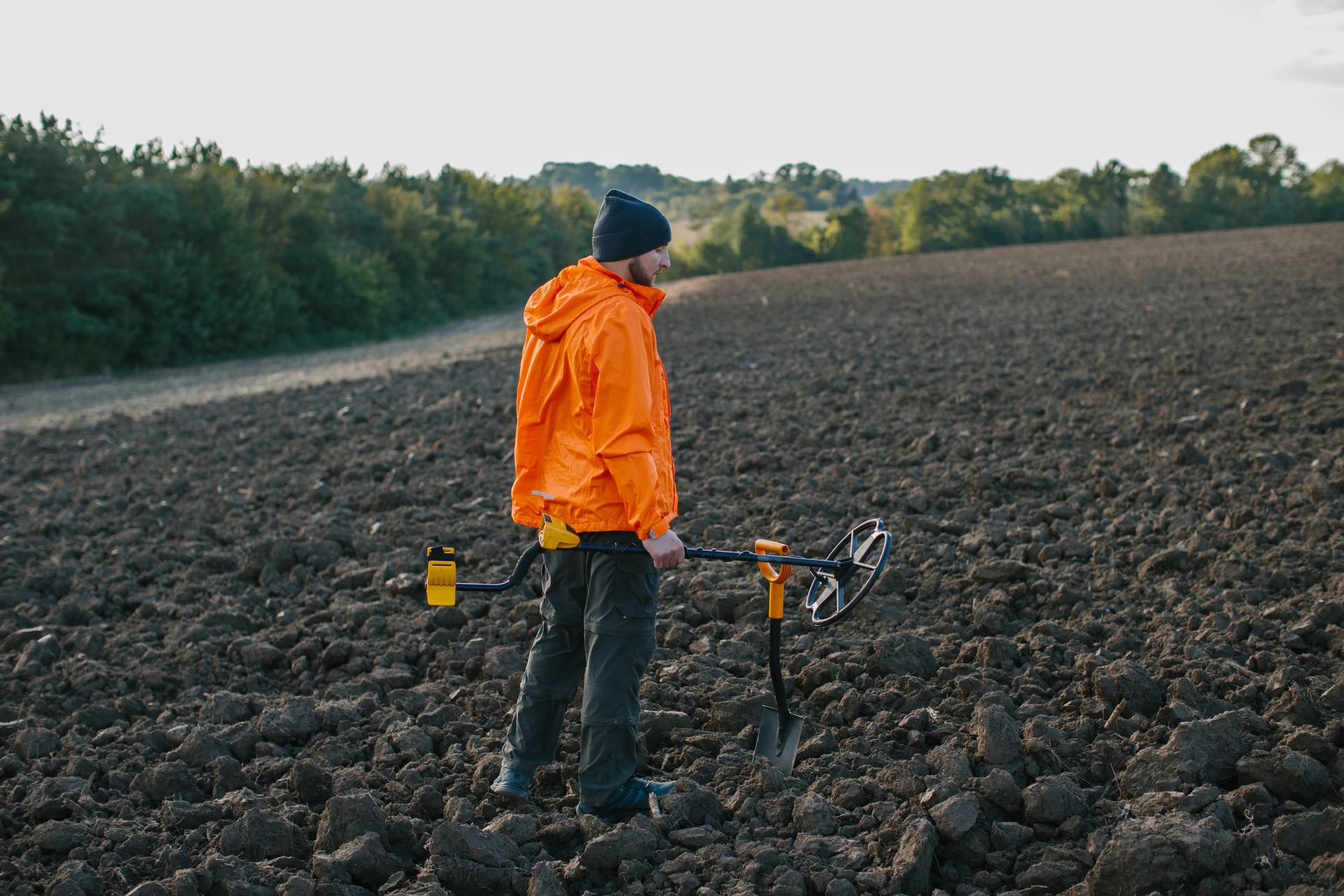 Person in orange jacket using a metal detector in a plowed field.