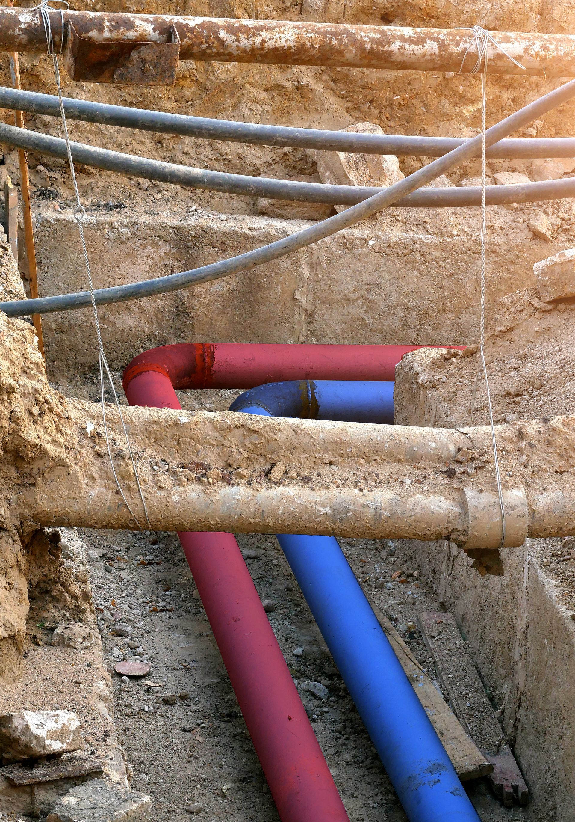 Pipes in red and blue in an open trench, alongside other utility lines, soil, and wooden supports.