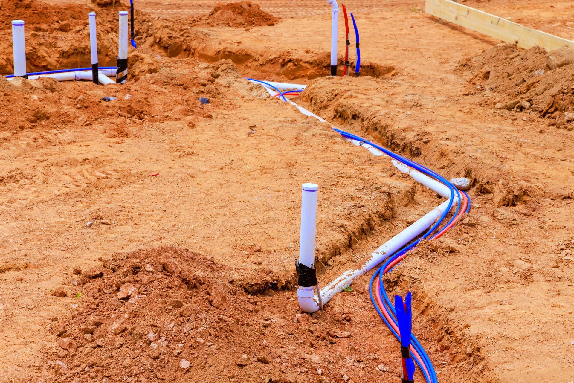 Pipes and plumbing are laid in trenches in the dirt at a construction site.