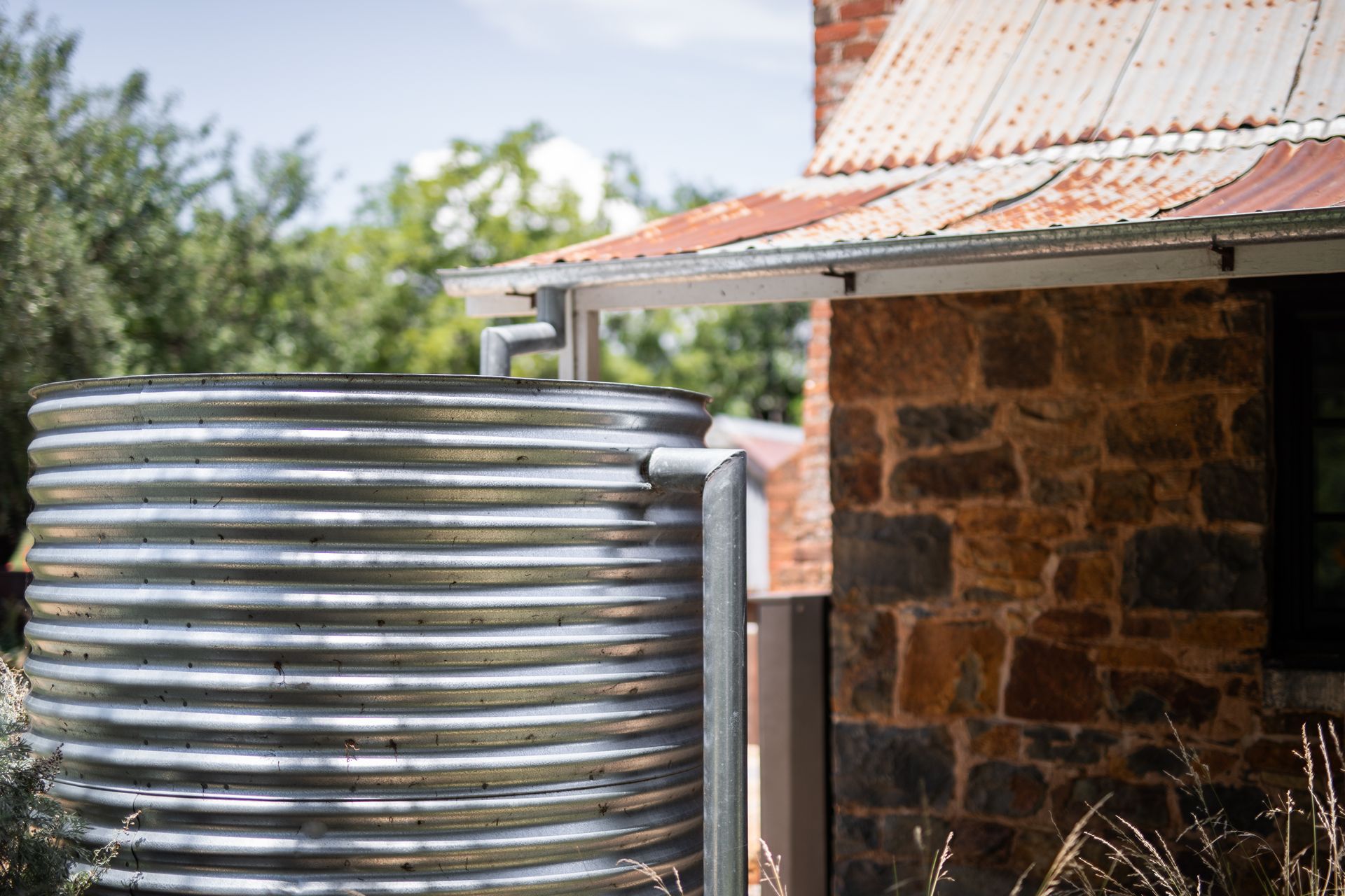 Corrugated metal water tank next to a stone building with a rusted metal roof; water flows from a gutter.