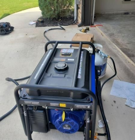 Blue generator with a black frame and propane tank outside a garage.