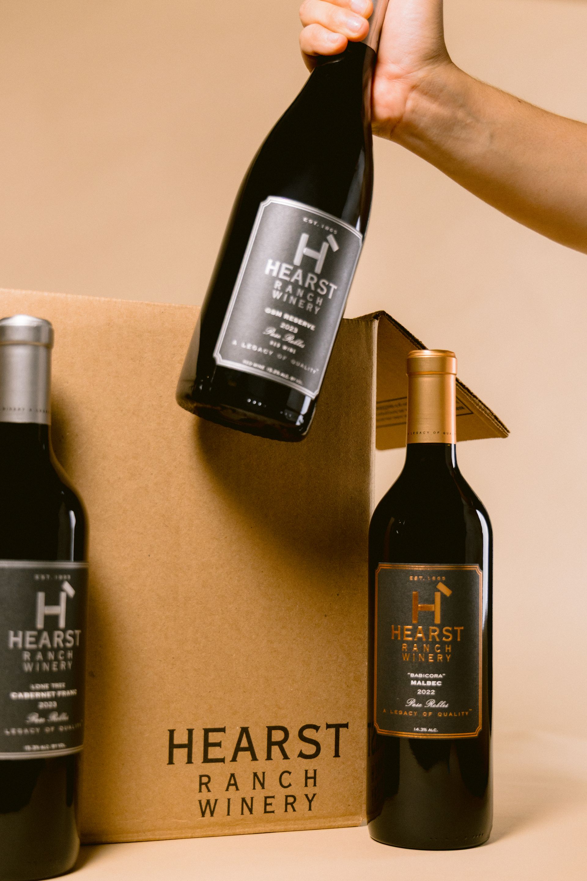 Hand holding a black wine bottle above two other wine bottles and a Hearst Ranch box on a tan background