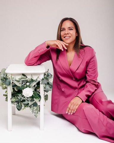 A woman in a pink suit is sitting next to a white stool with flowers on it.