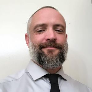 A man with a beard is wearing a white shirt and black tie.