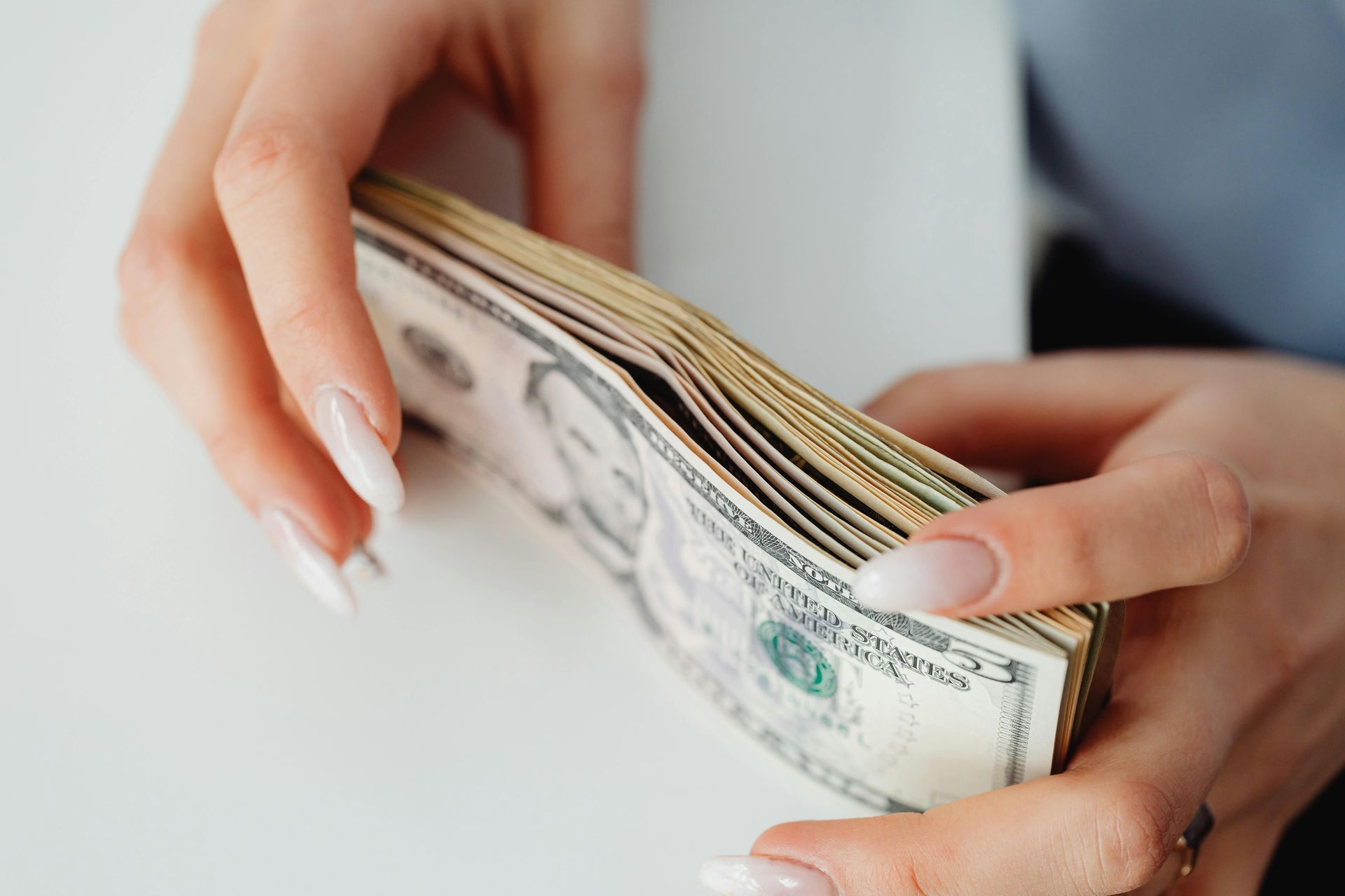 Hands hold a stack of US five-dollar bills against a white surface.