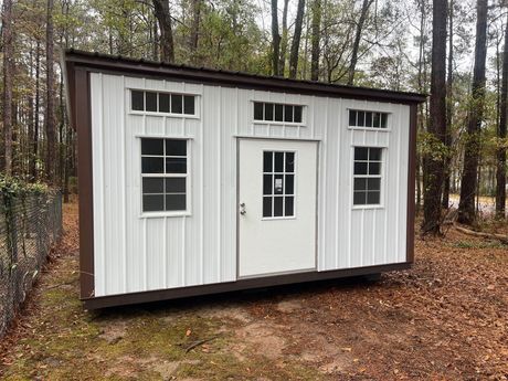 White shed with brown trim, door and windows, set in a wooded area.