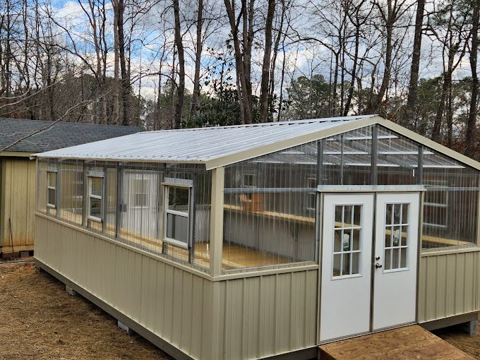 Greenhouse with beige walls, clear roof panels, and double white doors, set in a wooded area.