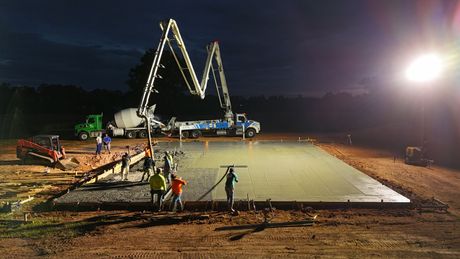 Concrete pour at night with trucks, pump arms, and workers leveling the surface.
