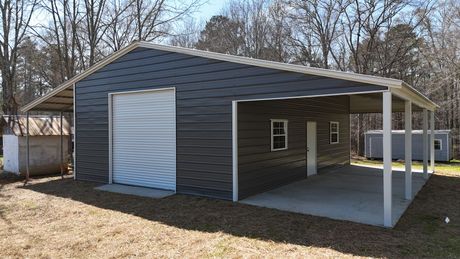 Dark gray metal workshop building with a roll-up door, side porch, and concrete slab, set in a wooded rural area.