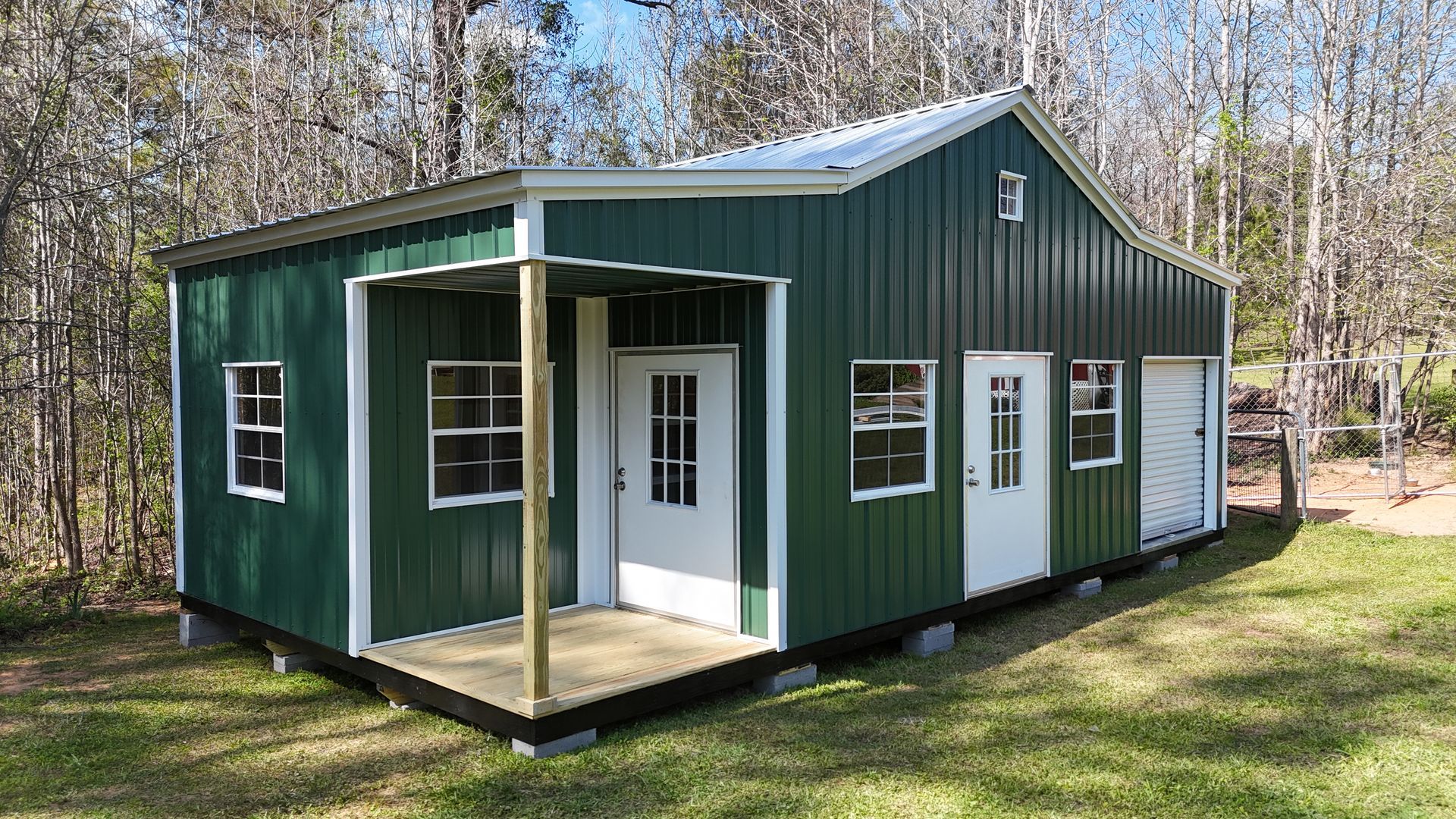 A green metal building with a small front porch, two white doors, several windows, and a small garage door in a wooded area.