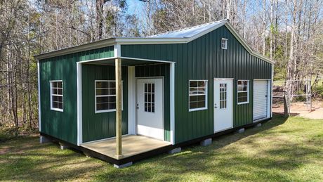 A green metal building with a small front porch, two white doors, several windows, and a small garage door in a wooded area.