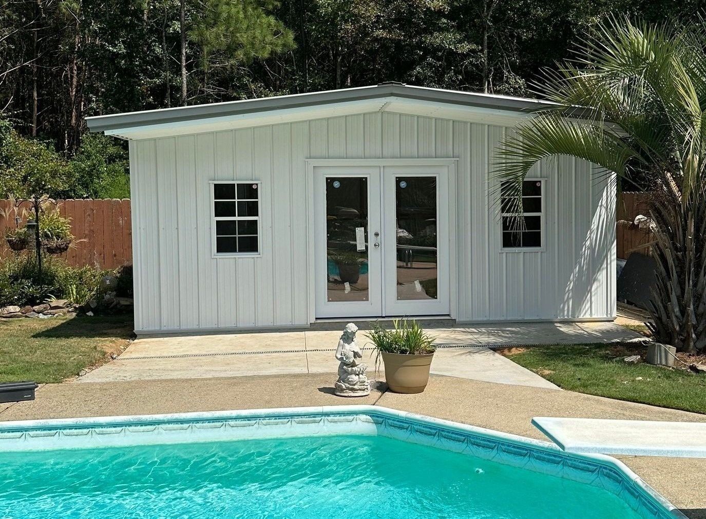 White shed with double doors and two windows, set in a yard with a palm tree and trees in the background.