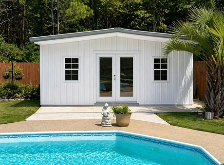 White shed with double doors and two windows, set in a yard with a palm tree and trees in the background.