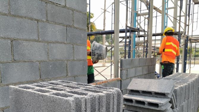 Construction workers in orange vests building a cinder block wall on a scaffold