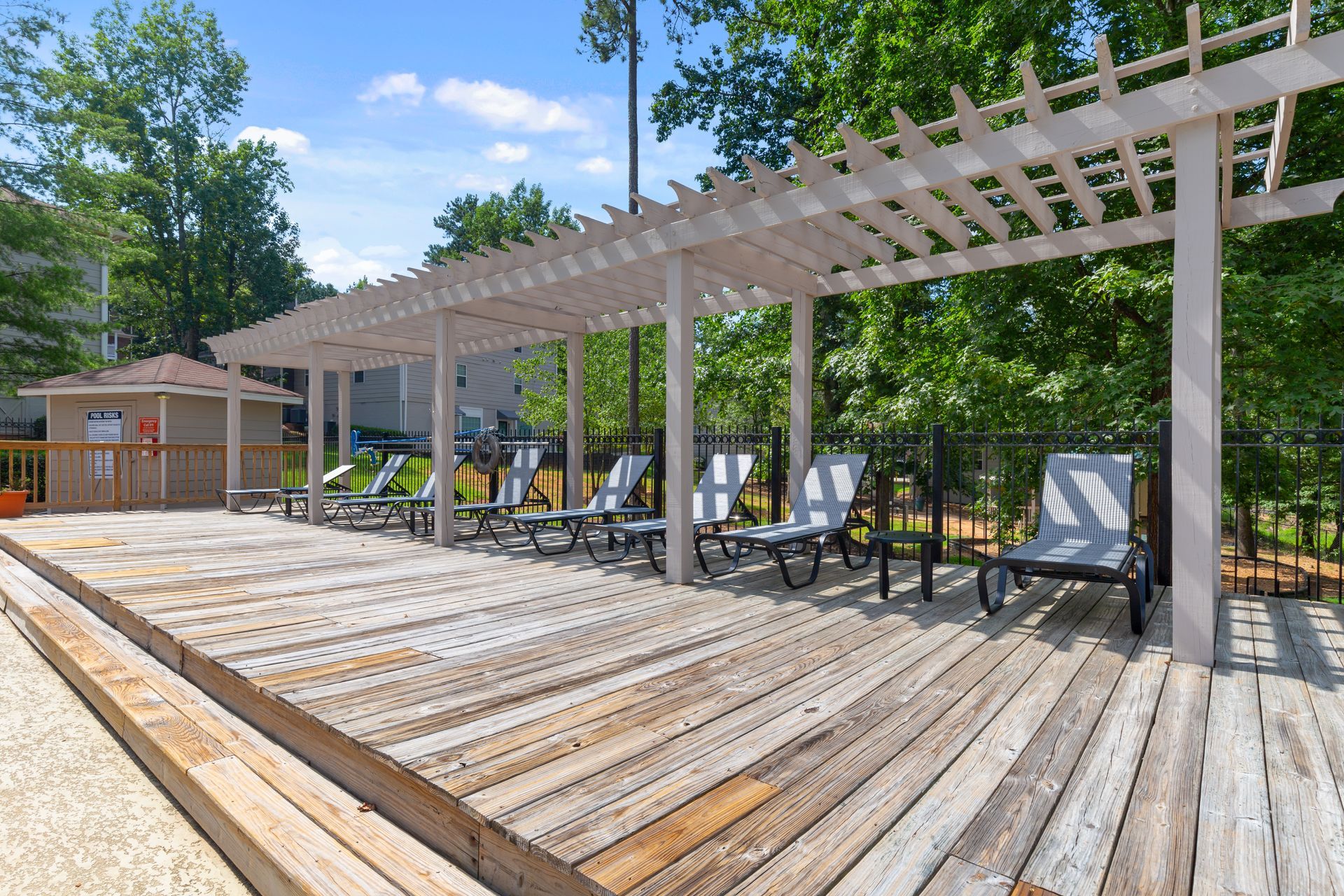 A row of lounge chairs on a wooden deck under a white pergola at an outdoor pool area with trees in the background.