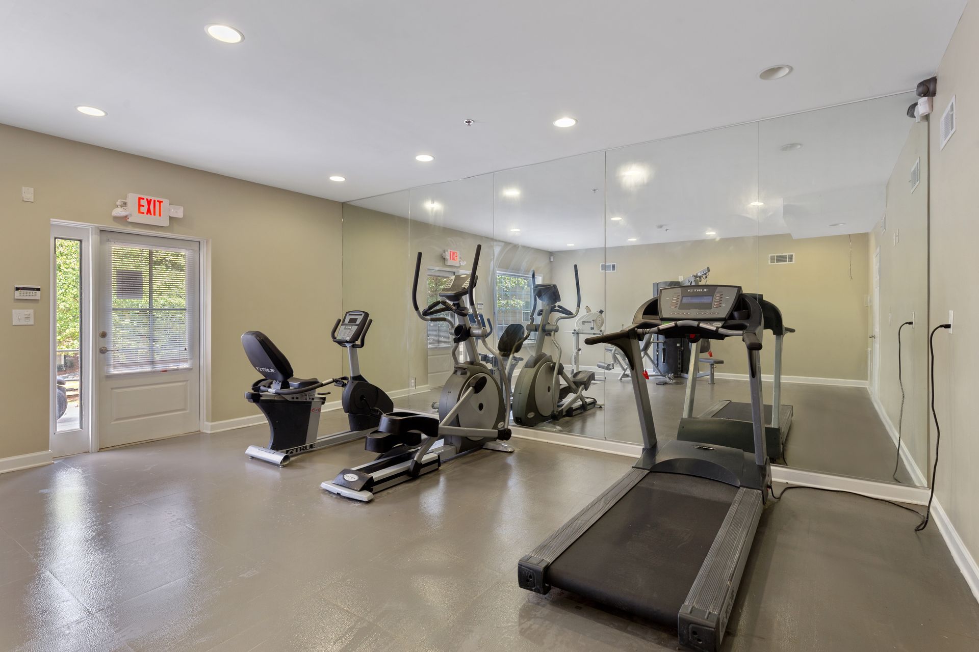 A fitness room with a treadmill, elliptical, and recumbent bike, featuring a large wall mirror and an exit door.